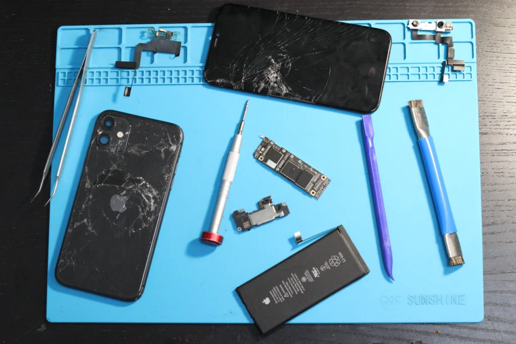 iPhone à l’écran fissuré démonté sur un tapis de réparation, avec batterie et outils